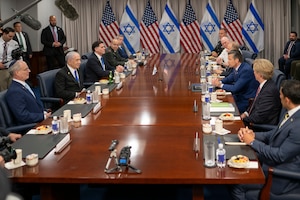 Nine men in business suits and two men in dress military uniforms are sitting at a large wooden table; there are five men on one side and six on the opposite side. There are water bottles and small snack dishes in front of them. In the background are the flags of the U.S. and Israel.