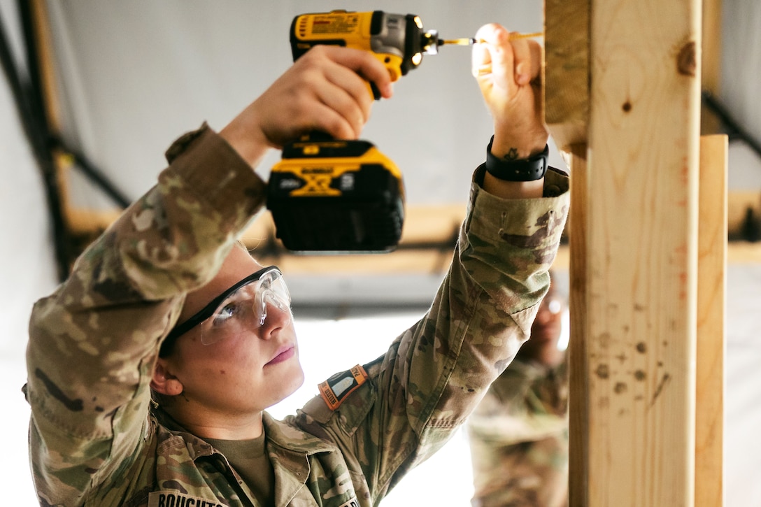 A soldier uses a cordless drill to apply a screw to a wood support beam as another soldier looks on in the background.