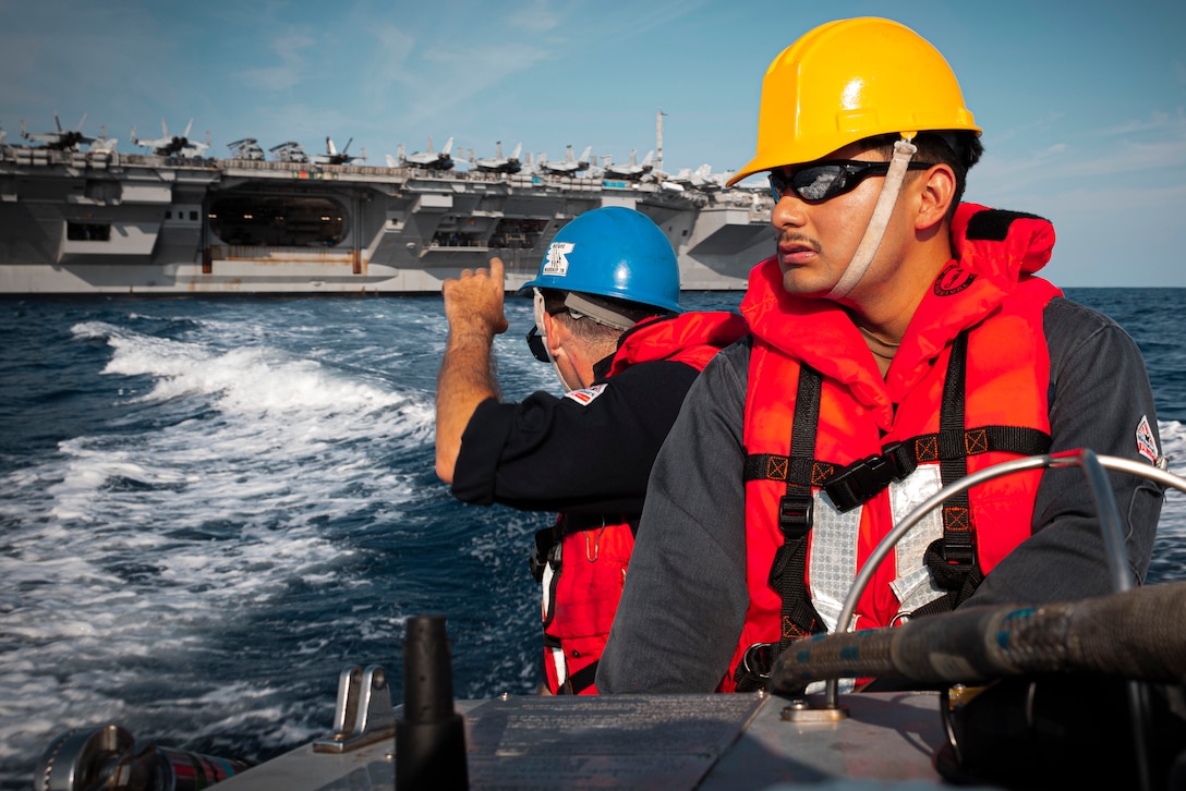 Two sailors, one looking back toward a nearby aircraft carrier, operate a small boat in the open ocean on a sunny day.