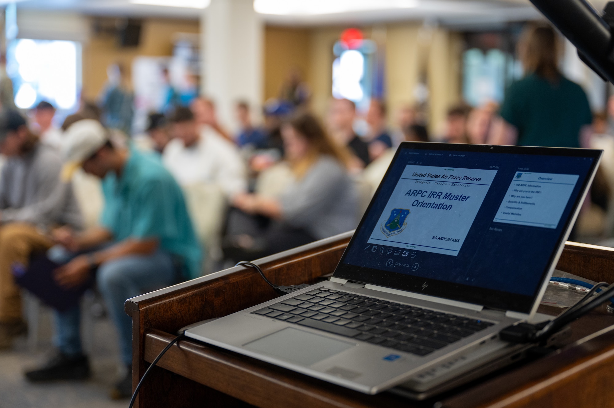 a laptop computer sits open with a slide show presentation on the screen, in front of a room filled with people. this photo signifies that a presentation is being given.