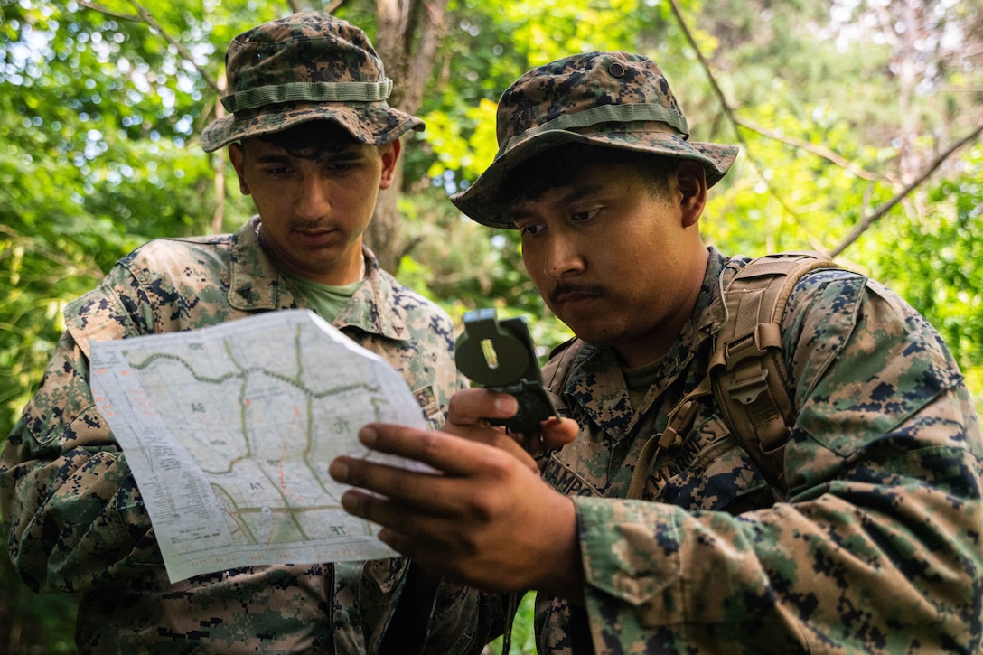 Two Marines wearing floppy hats use a compass and a map in a wooded area on a sunny day.