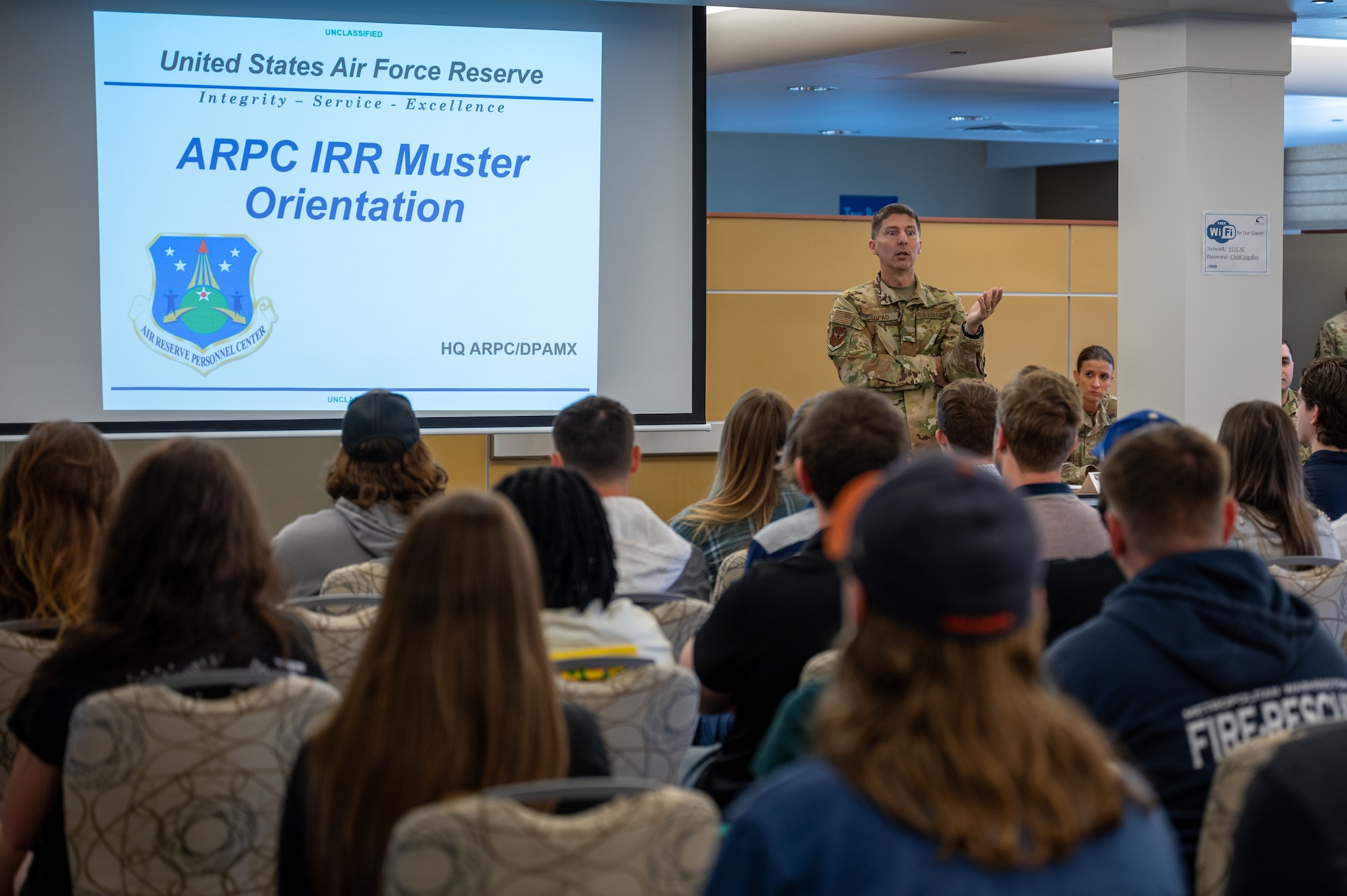 a man in Air Force Camouflage speaks to a room of people in civilian attire