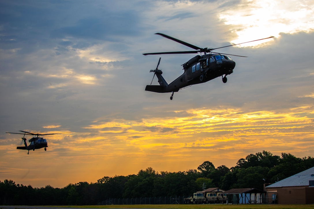 Soldiers operate two Black Hawk helicopters above an airfield, with an orange, cloudy sky, trees and a structure in the background.