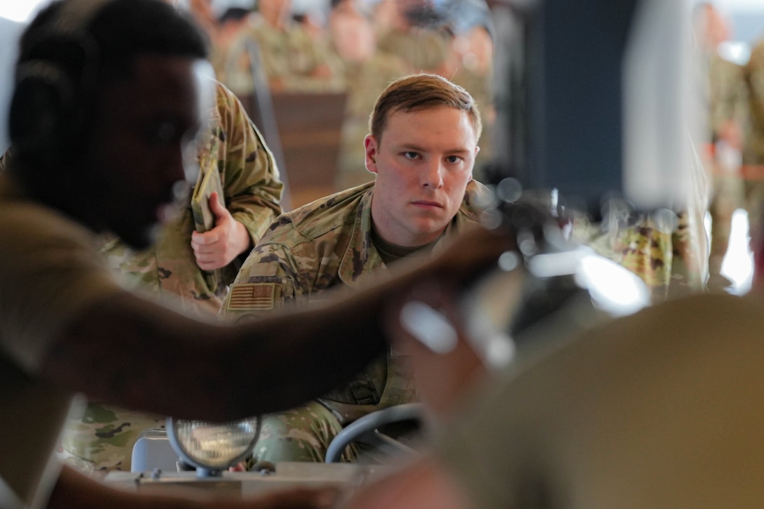 An airman watches other airmen work on an unseen device as others stand in the background.