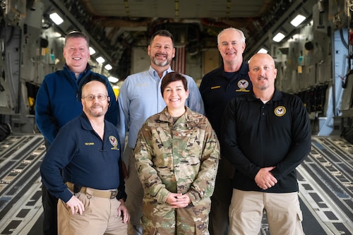 Five men in civilian attire pose for a photo with a woman in air force camouflage while aboard