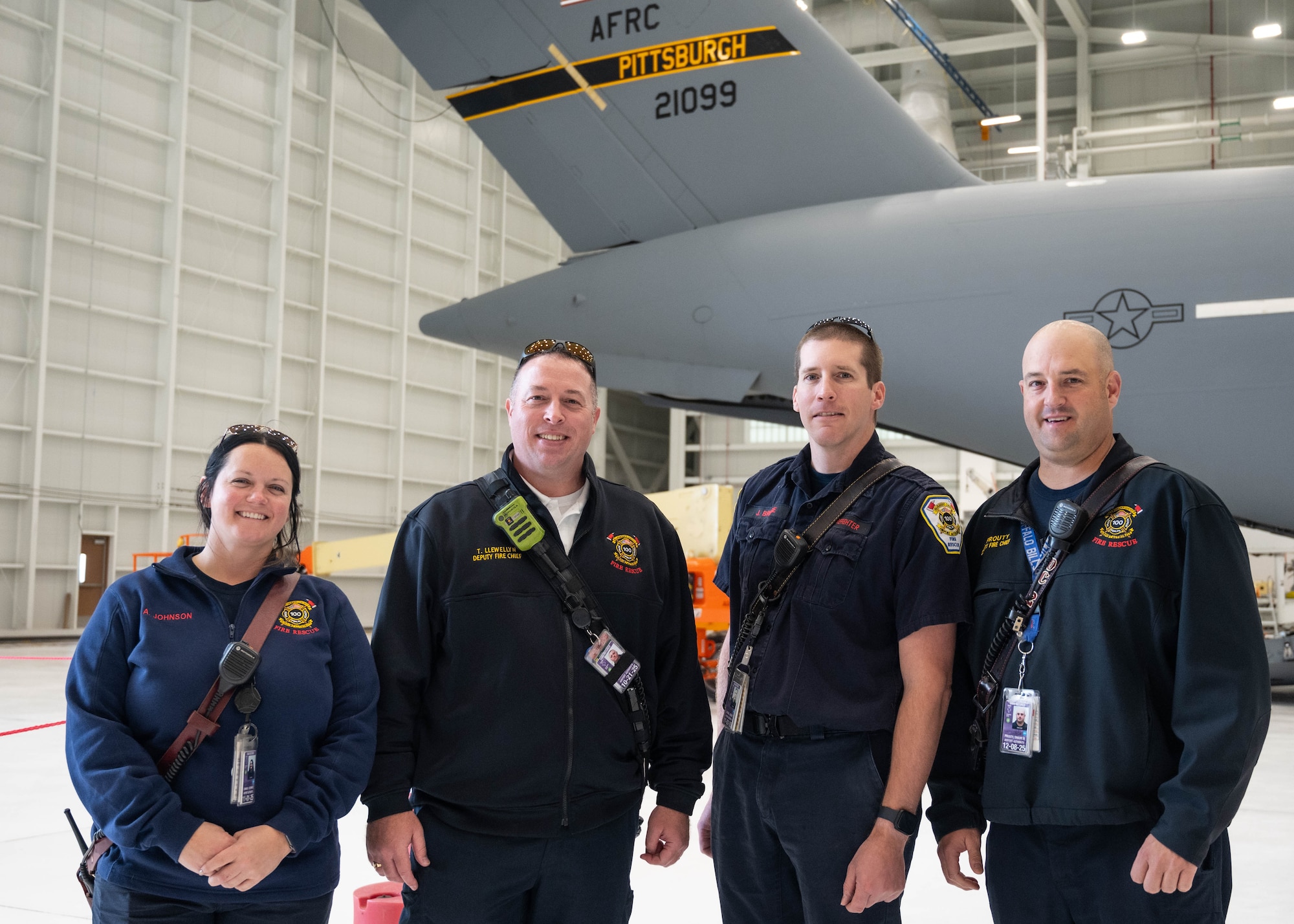 four fire fighters dressed in blue fatigues pose for a photo in an airplane hangar
