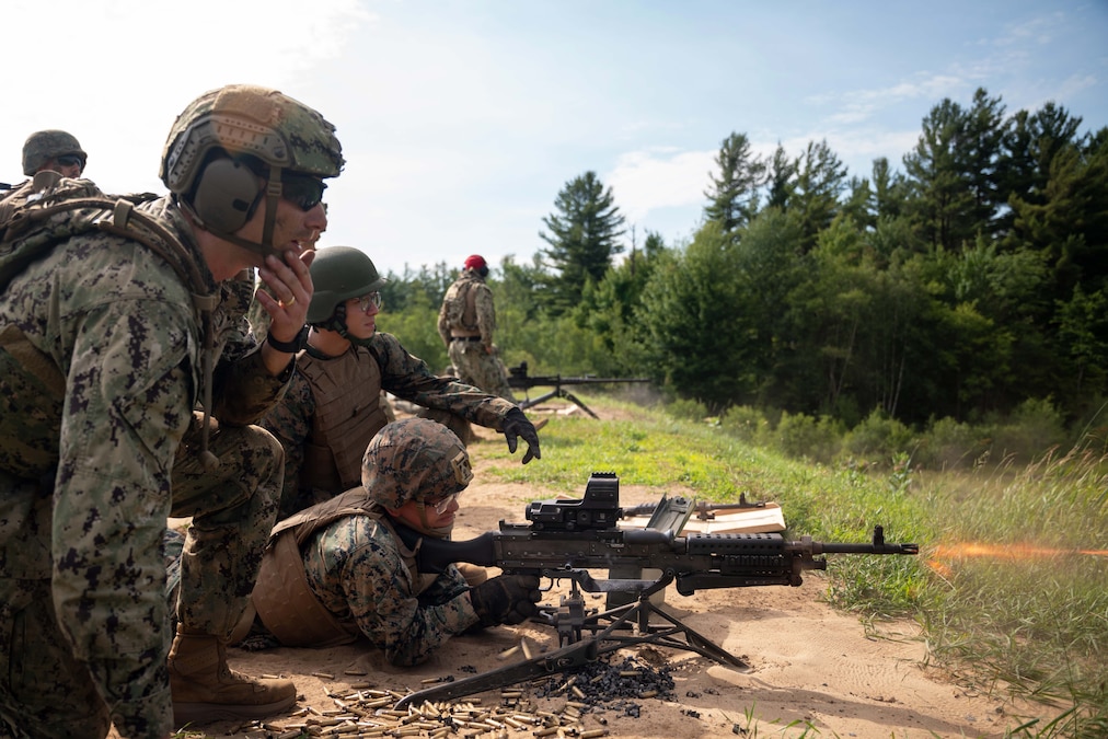 A group of Coast Guardsmen and Marines watch another one fire a machine gun from a lying position on a dirt and grassy area, with another machine gun and trees in the background.