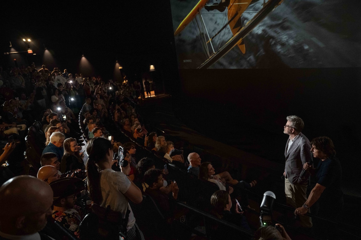 Two people stand in front of a movie screen with a crowd of people watching from their seats.