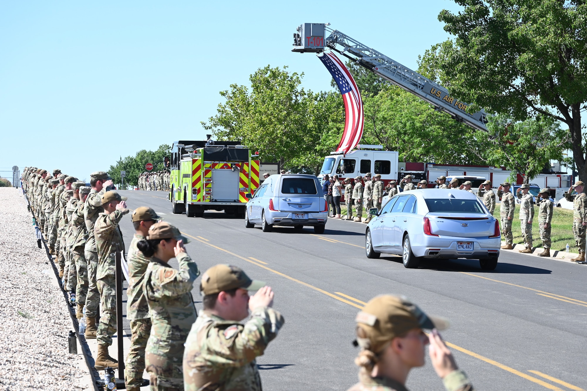 Airmen lining a road salute a procession of vehicles.