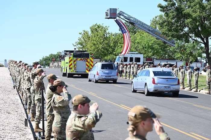 Airmen lining a road salute a procession of vehicles.