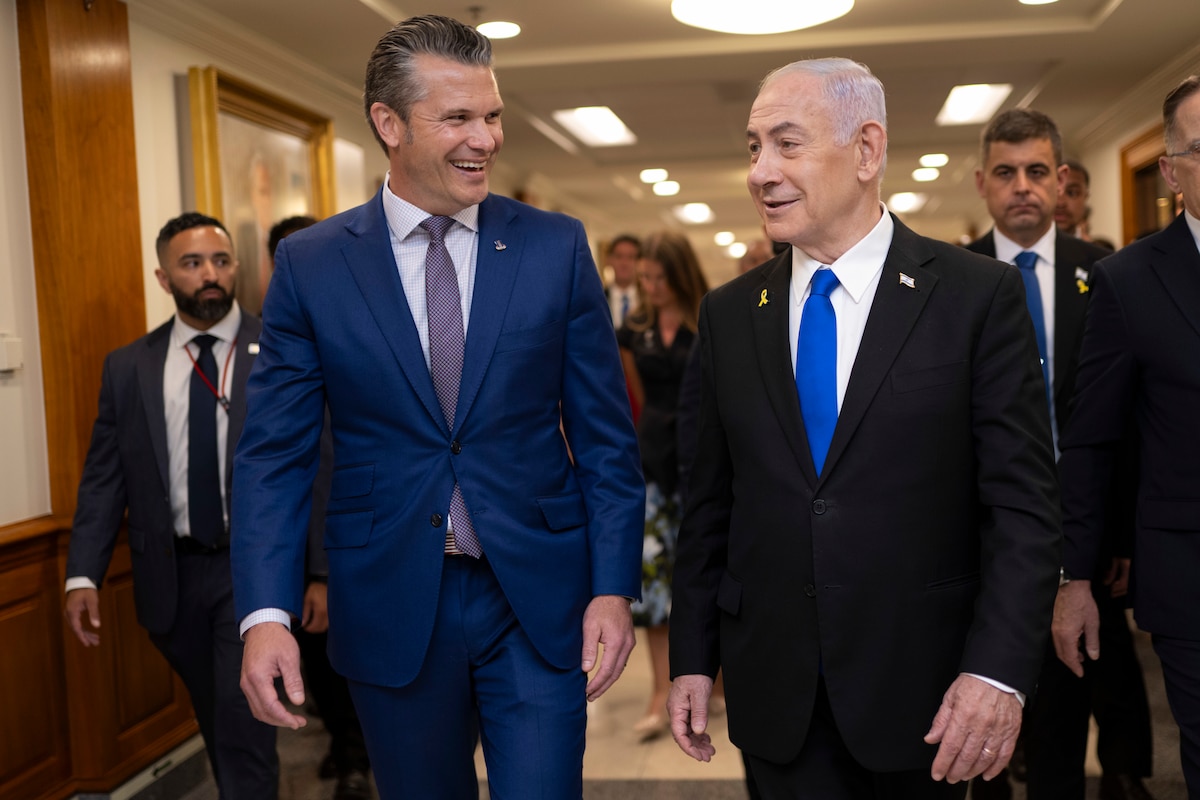 Two men in business suits are smiling as they walk and talk in a large hallway. There are three additional men in business suits walking behind them in the background.