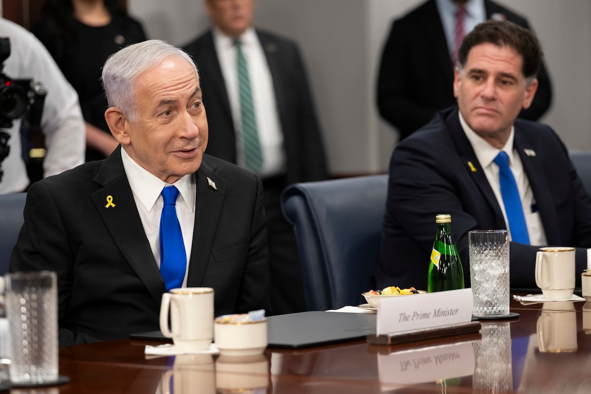 A man wearing a business suit is sitting at a wooden table, looking to the right of the screen. A sign resting on the table in front of him reads “The Prime Minister.” There is a second man, also in a business suit, in the background who is looking at the first man.
