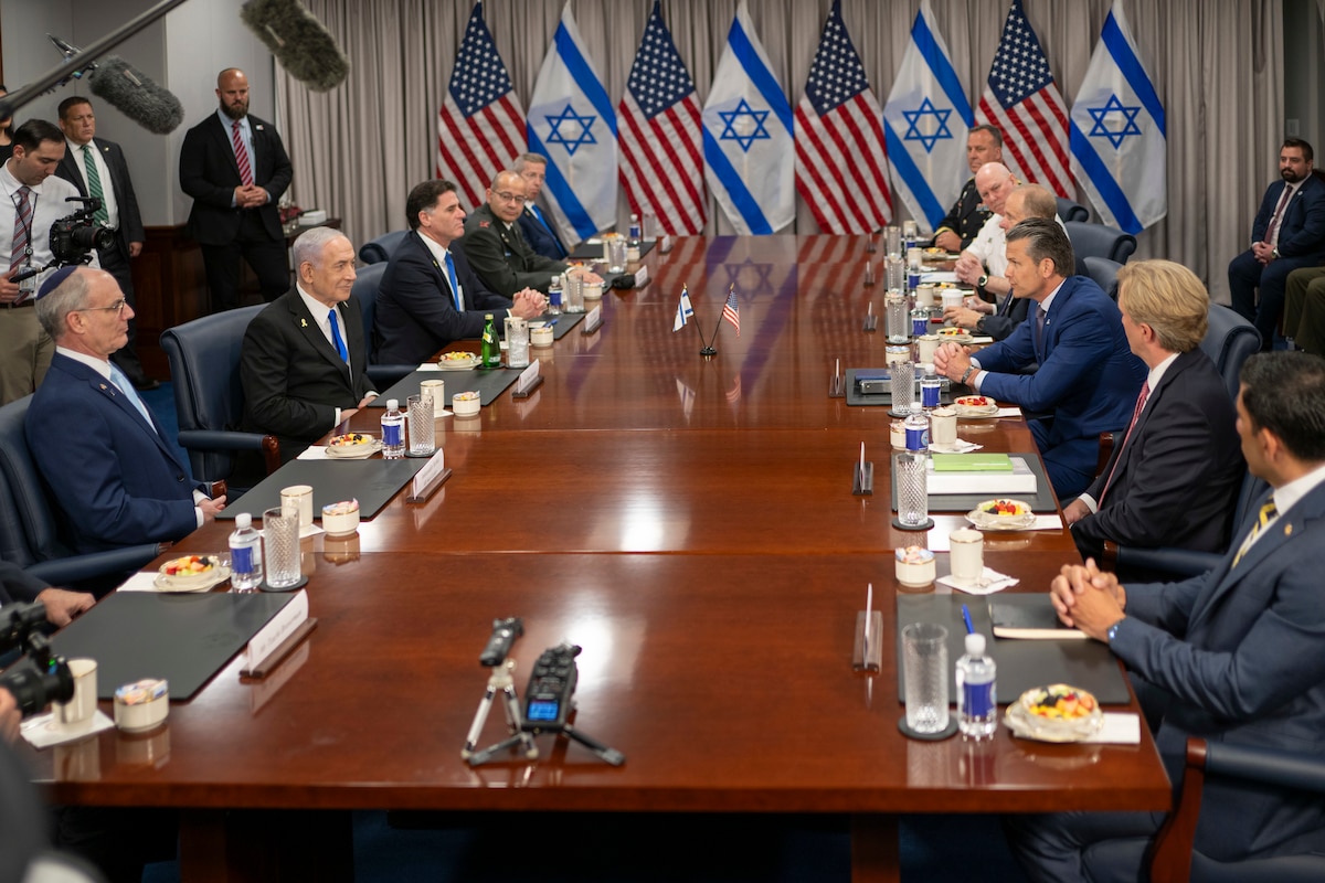 Nine men in business suits and two men in dress military uniforms are sitting at a large wooden table; there are five men on one side and six on the opposite side. There are water bottles and small snack dishes in front of them. In the background are the flags of the U.S. and Israel.