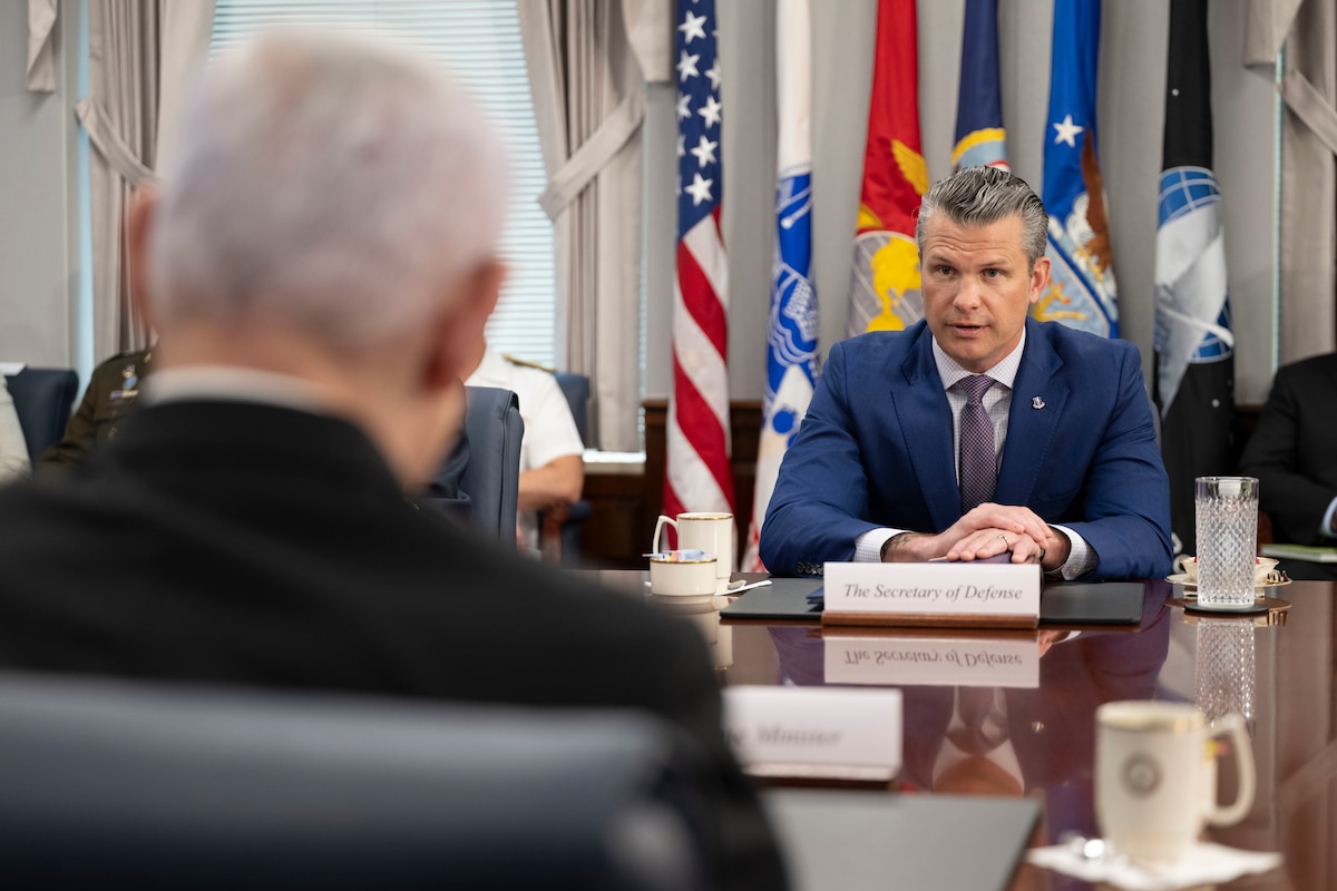 A man in a business suit is sitting on one side of a wooden table, speaking to another man in a business suit with his back to the camera. A small sign reading “Secretary of Defense” is in front of the first man, and there is a group of rolled-up flags behind him.