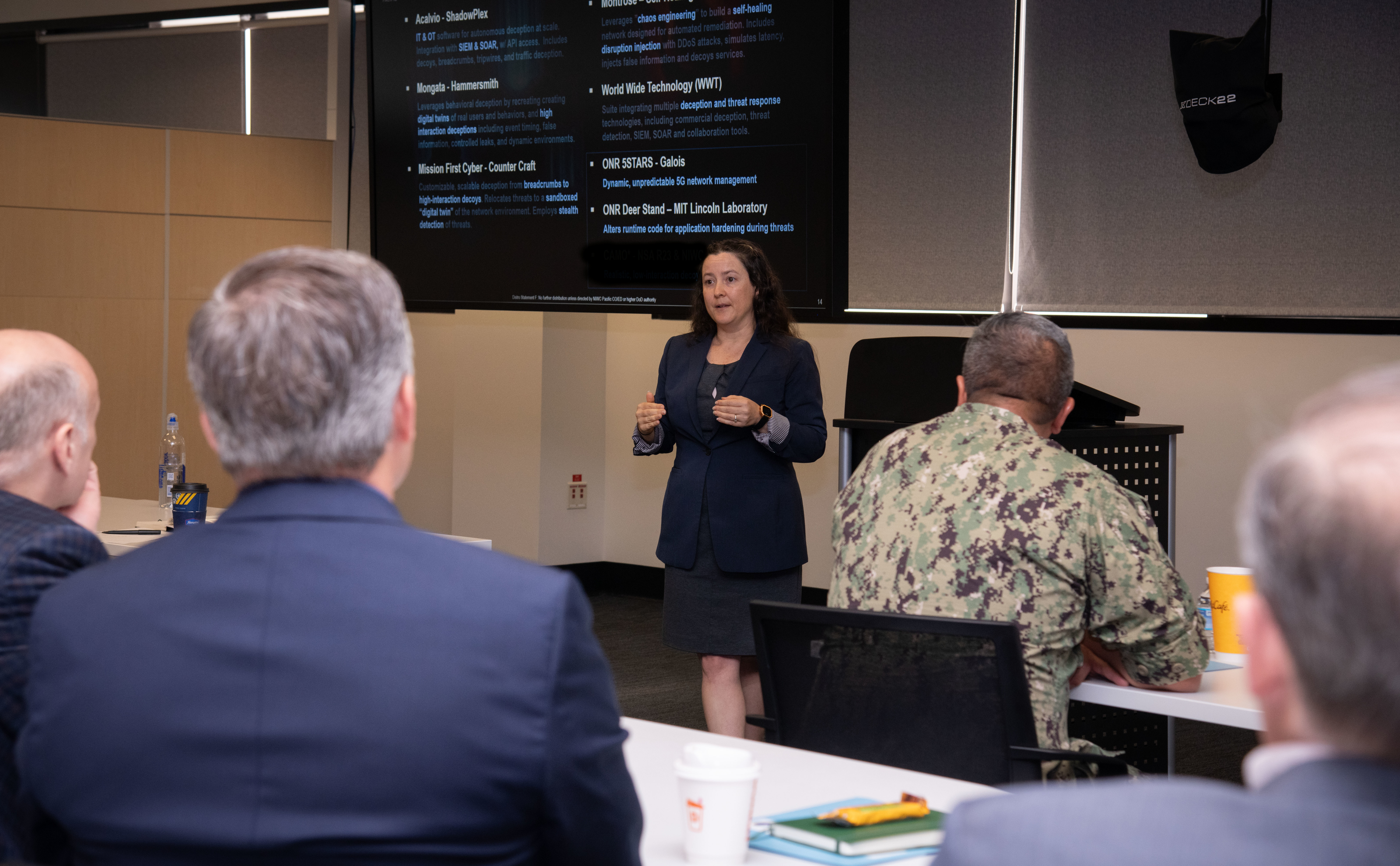 Photo of NIWC Pacific and Atlantic workforce at CyberANTX having a brief discussion.  Woman in black in front of suited men and one male navy seaman in camouflage uniform.