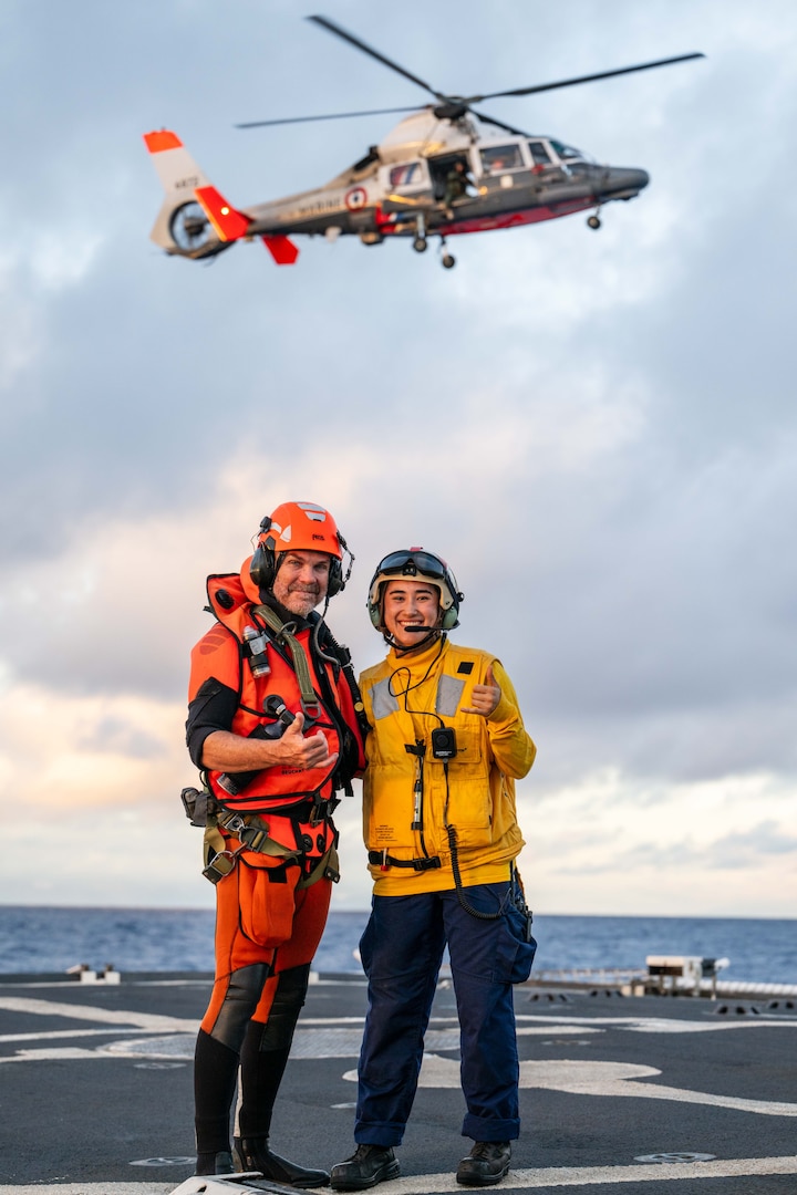 Ensign Junna Castel, Landing Signal Officer for U.S. Coast Guard Cutter Harriet Lane (WMEC 903) poses for a quick photo with a Marine Nationale rescue swimmer while a French Navy Flottille 35F AS365 Dauphin helicopter hovers above the flight deck during joint flight operations off the coast of Tahiti, French Polynesia, May 31, 2025. Presence and human-to-human interaction builds the connective tissue that strengthens the safety and security of all countries and fosters a committed network of partners with shared principles and norms in the maritime domain. (U.S. Coast Guard photo by Petty Officer 3rd Class Austin Wiley)