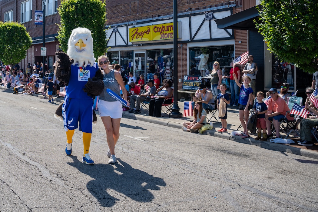 A bald eagle mascot wearing blue athletic gear walks alongside a tall blonde woman in grey tank top and white shorts, crowd of people waving U.S. flags can bee seen int he background