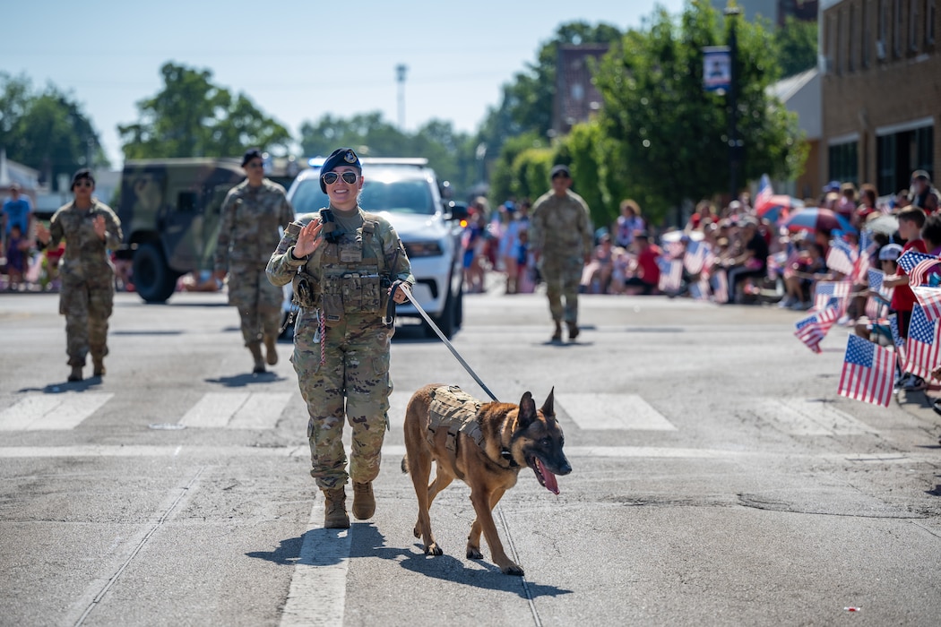 Sgt. Swisher, wearing camouflage military uniform, waves as she holds the leash of a German Shepperd