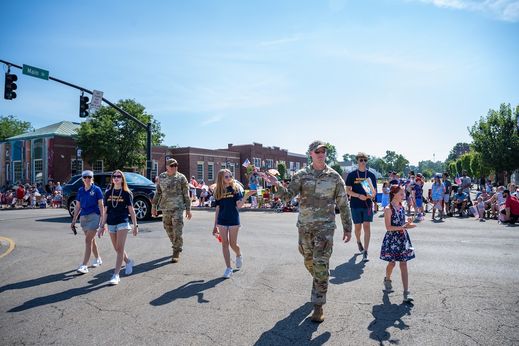 A group of people, some in military camouflage and other in civilian attire, march through the street waving and smiling