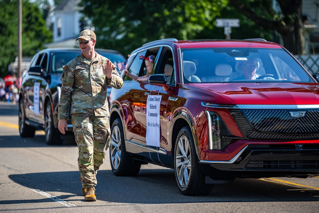 Col. Richards, wearing camouflage military uniform, waves while walking beside a red SUV with his daughter waving from the window