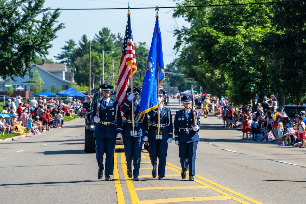 Four Air Force service members, dressed in blue service dress uniforms, march towards viewer carrying ceremonial rifles and U.S. and Air Force flags. Crowds of people can be seen on either side of photo
