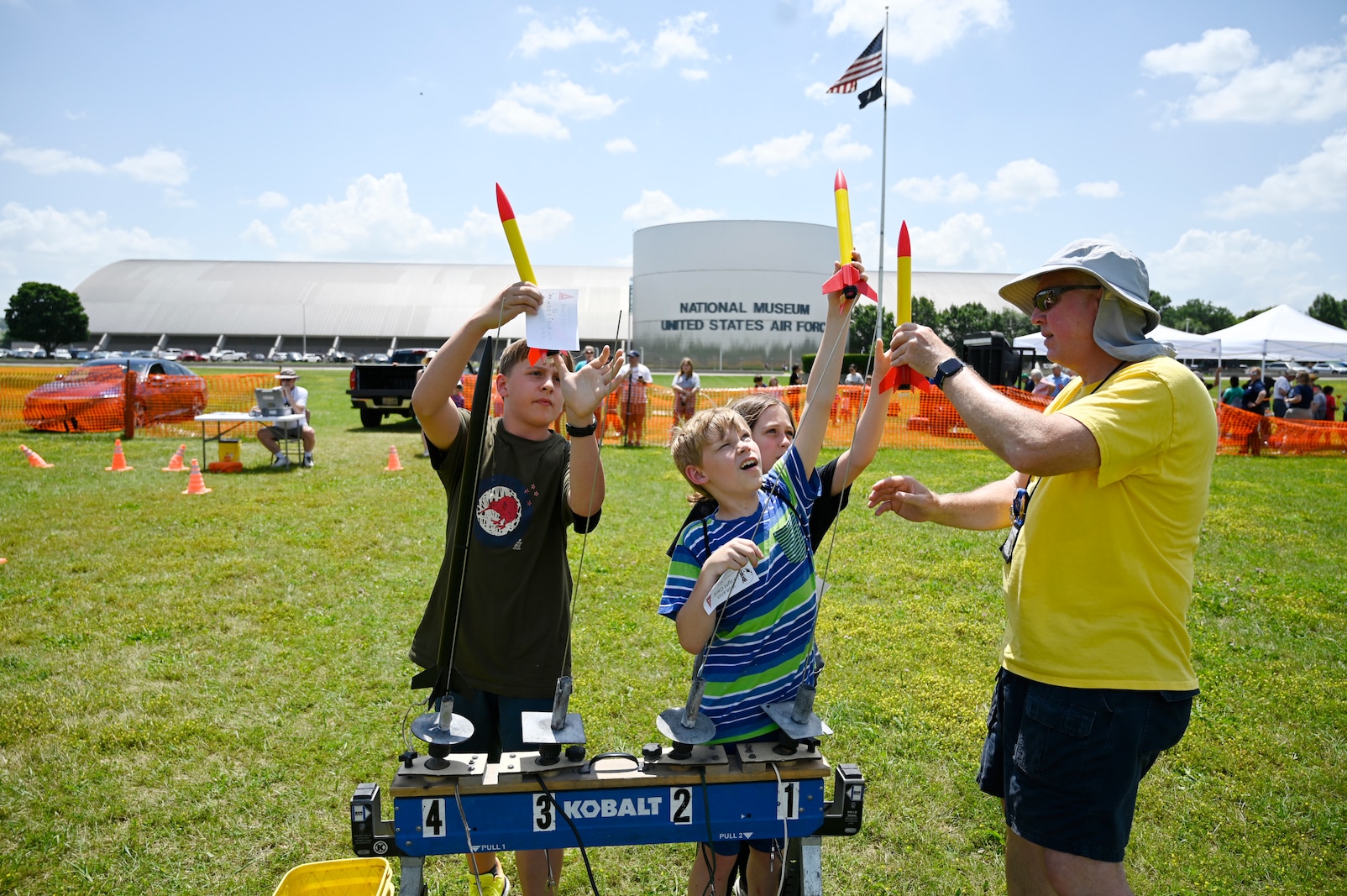 A volunteer instructor holds a model rocket while explaining the launch process to a group of students during Aerospace Camp at the National Museum of the U.S. Air Force. The students watch closely, gathered around the instructor outdoors on the museum grounds.