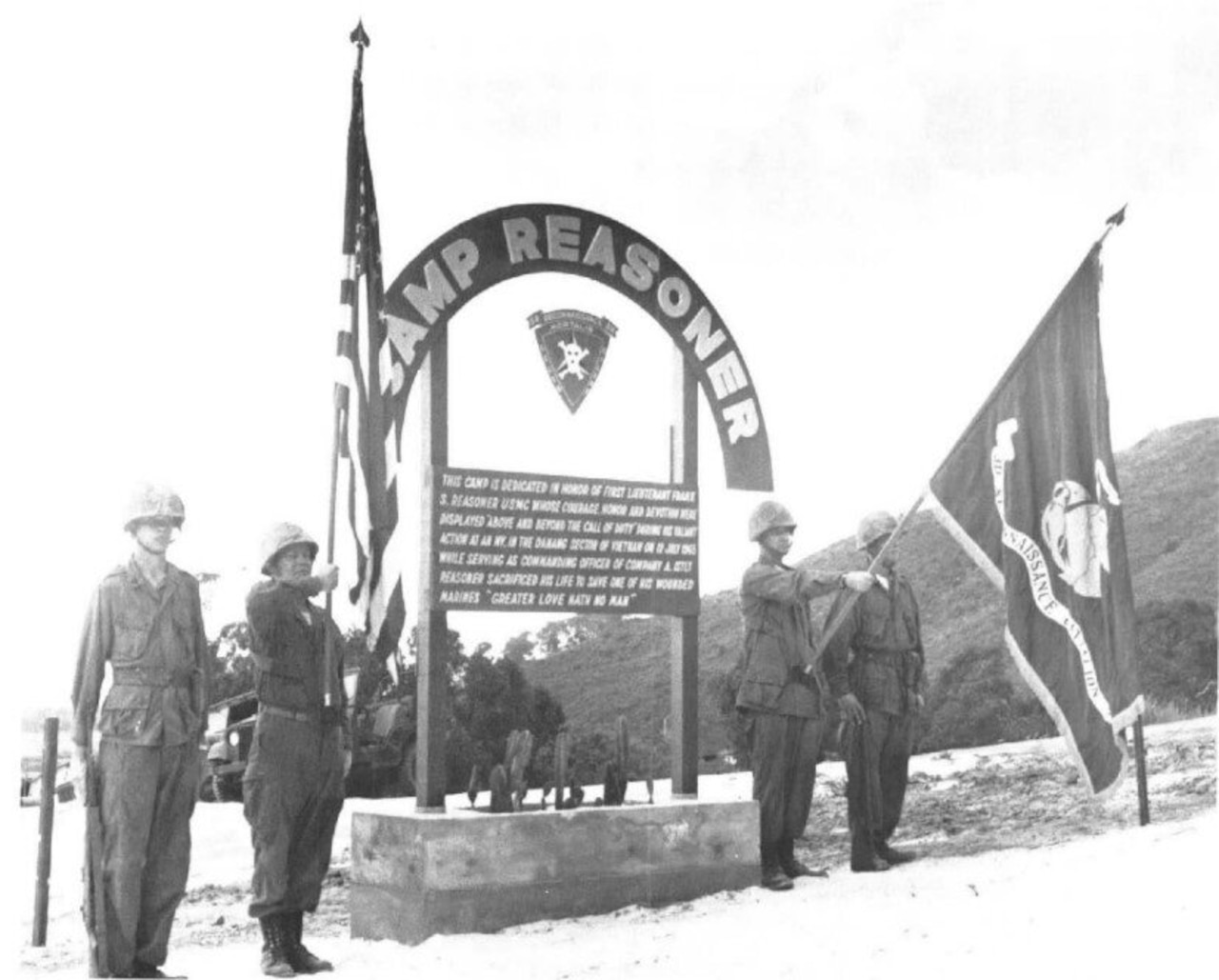 Four men wearing military uniforms and helmets stand in front of a sign that reads “Camp Reasoner.” One man is holding the American flag, while another holds the Marine Corps flag.