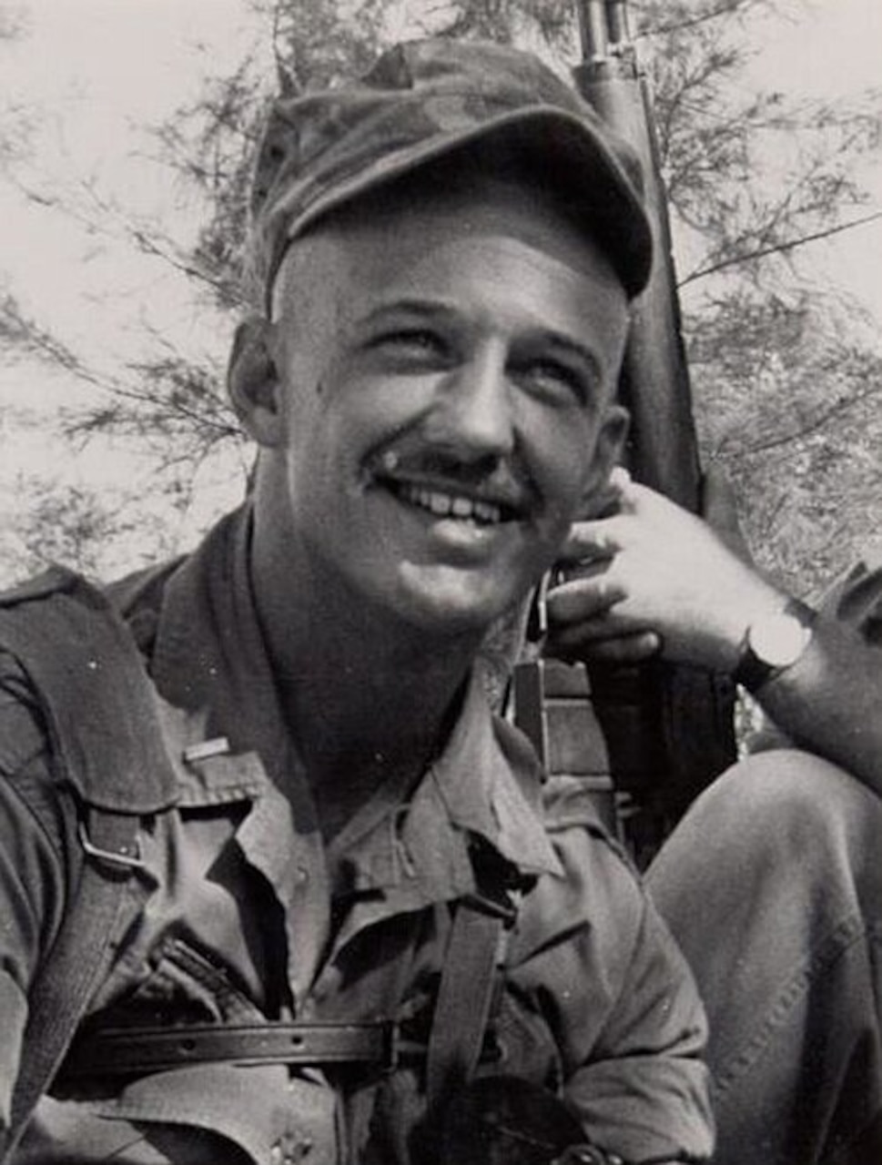 A man in a military combat uniform smiles. Another man in a military uniform holding a gun sits behind him.