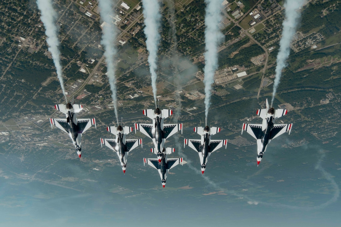 Six aircraft fly in formation above a green and tan landscape, as seen from upside down.