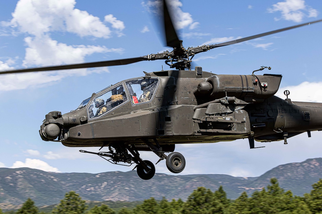 A helicopter flies above trees with mountains and blue skies with clouds in the distance.