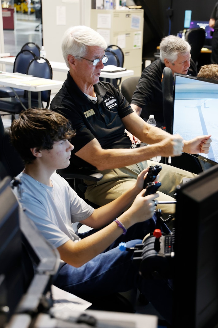 A student sits at a flight simulator station with a large screen showing an in-flight view, hands on the controls, while a volunteer instructor beside them offers guidance. Other simulator stations with students and instructors are visible in the background, showing a full classroom engaged in pilot training.