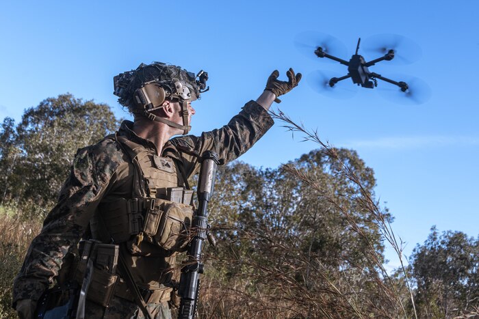 A Marine in tactical gear reaches toward a drone flying above a forest setting during the day.