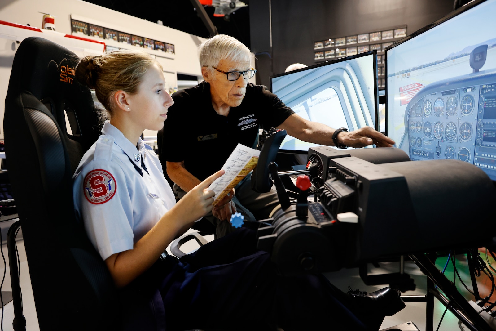A student sits at a flight simulator station with a large screen showing an in-flight view, hands on the controls, while a volunteer instructor beside them offers guidance.