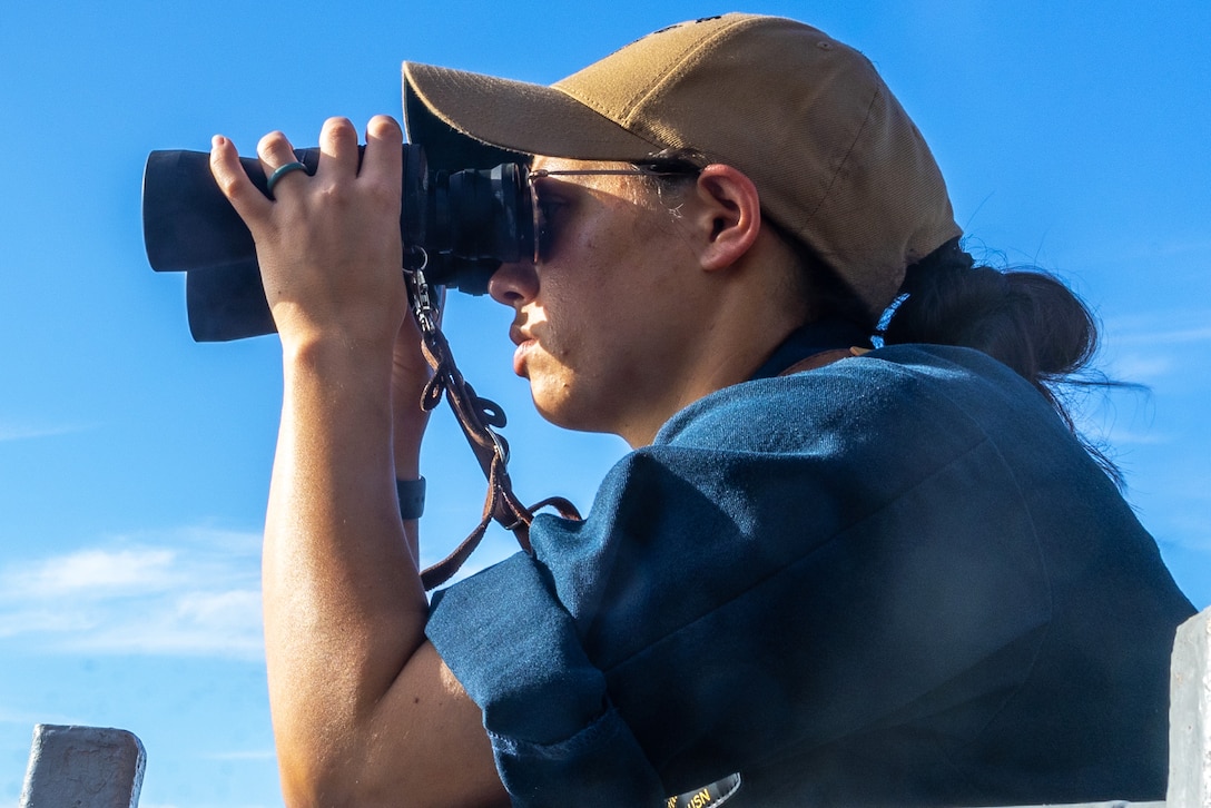 A sailor wearing a cap, sunglasses and blue shirt peers through binoculars on a sunny day.