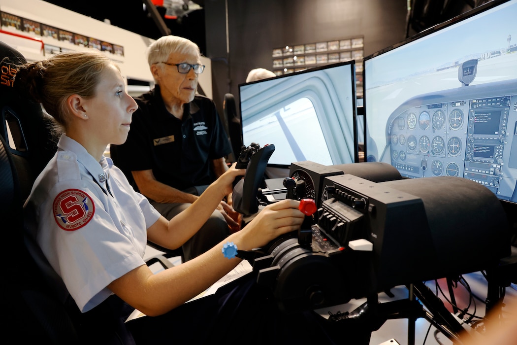 A student sits at a flight simulator station with a large screen showing an in-flight view, hands on the controls, while a volunteer instructor beside them offers guidance.