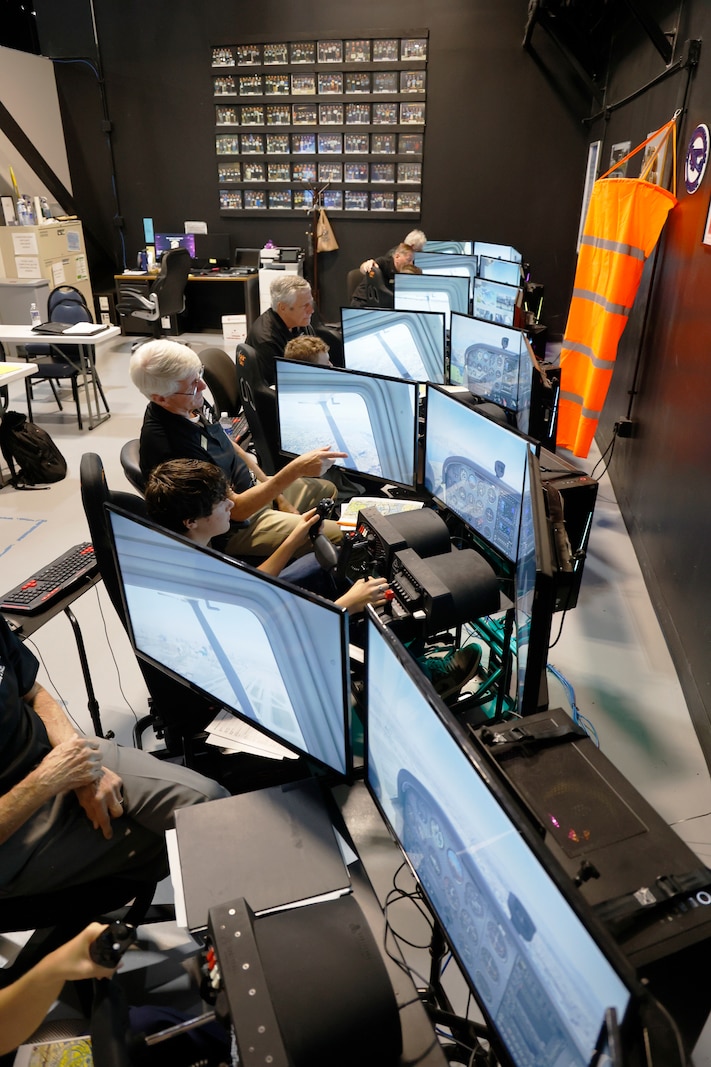 Students sit at a flight simulator stations with large screens showing an in-flight view, hands on the controls, while volunteer instructors beside them offers guidance.