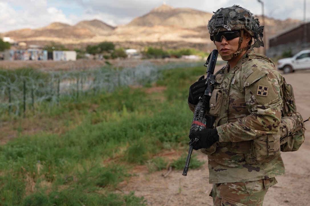 A soldier in combat gear stands on a dirt road, with grass, rows of concertina wire, structures and a truck in the background.
