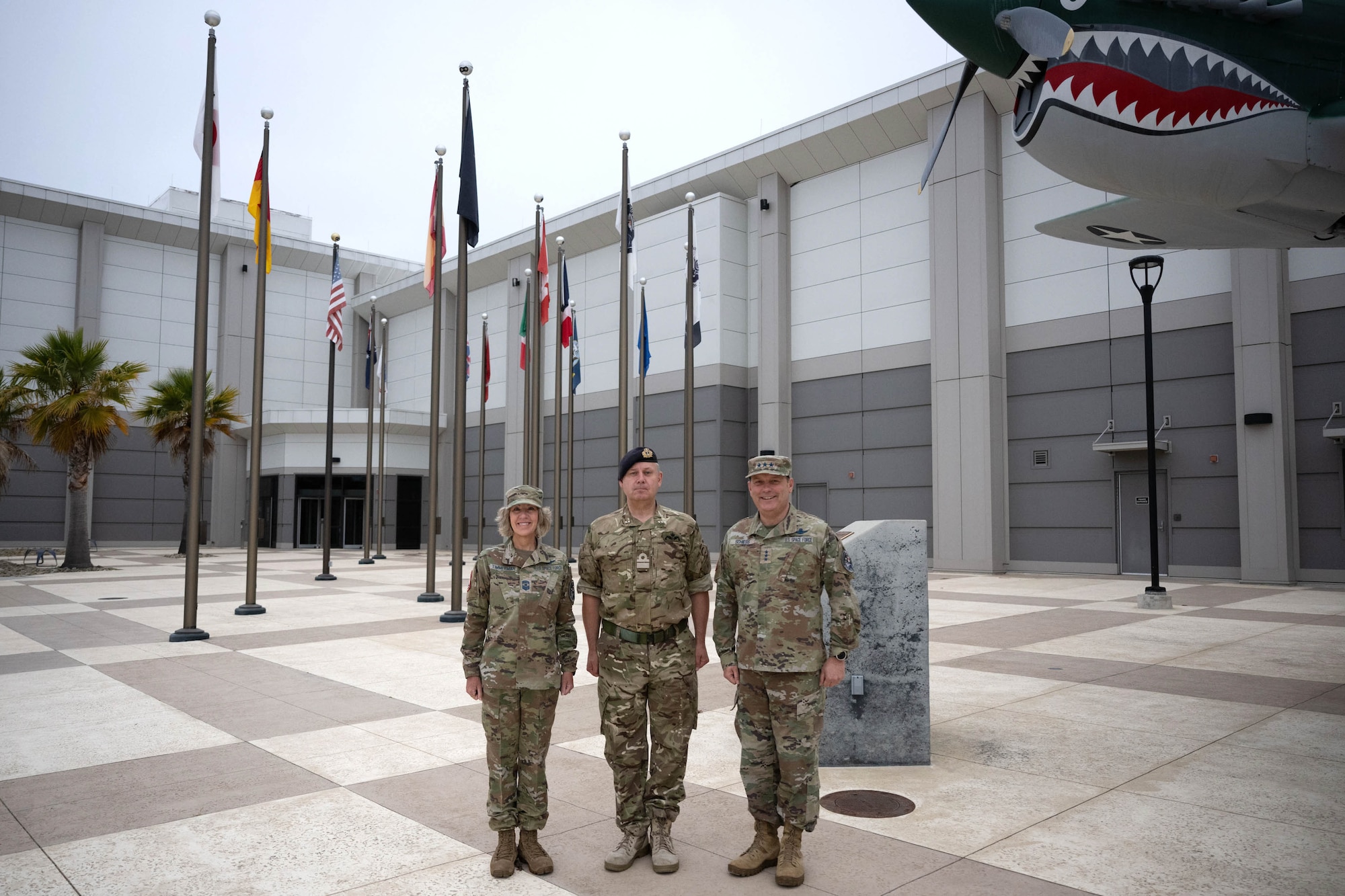 Two US and one UK military member stand in front of a building.