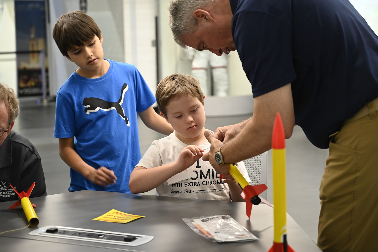 A student concentrates as he builds a model rocket with the guidance of a volunteer instructor. Another student nearby watches the process with interest. The activity takes place during the Modified Aerospace Camp at the National Museum of the U.S. Air Force.