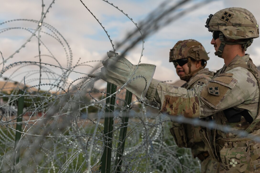 Two soldiers wearing protective gloves work on rows of concertina wire wrapped around stakes in a grassy area, with a hill in the background.