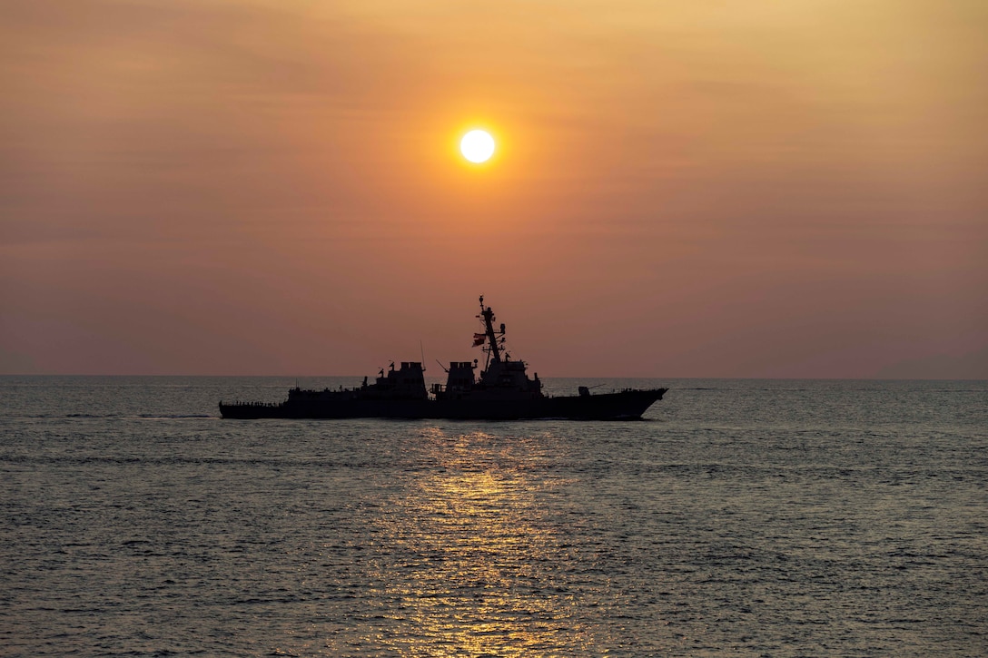 The silhouette of a ship sails on the ocean under an orange sun and sky.