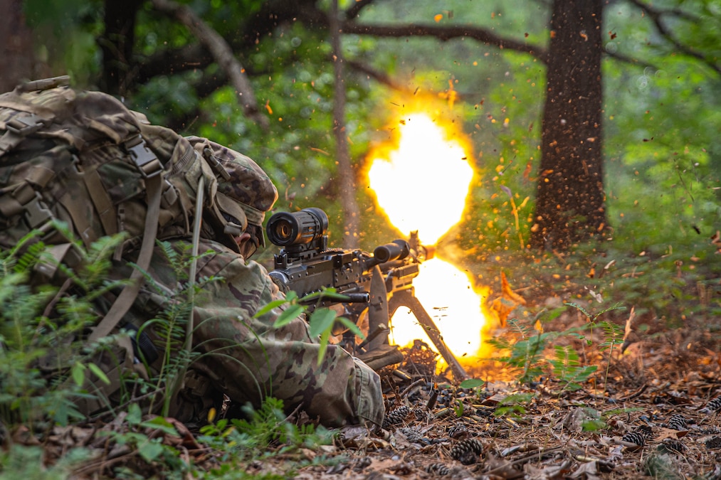 A soldier in tactical gear fires a gun while laying in a forest setting.