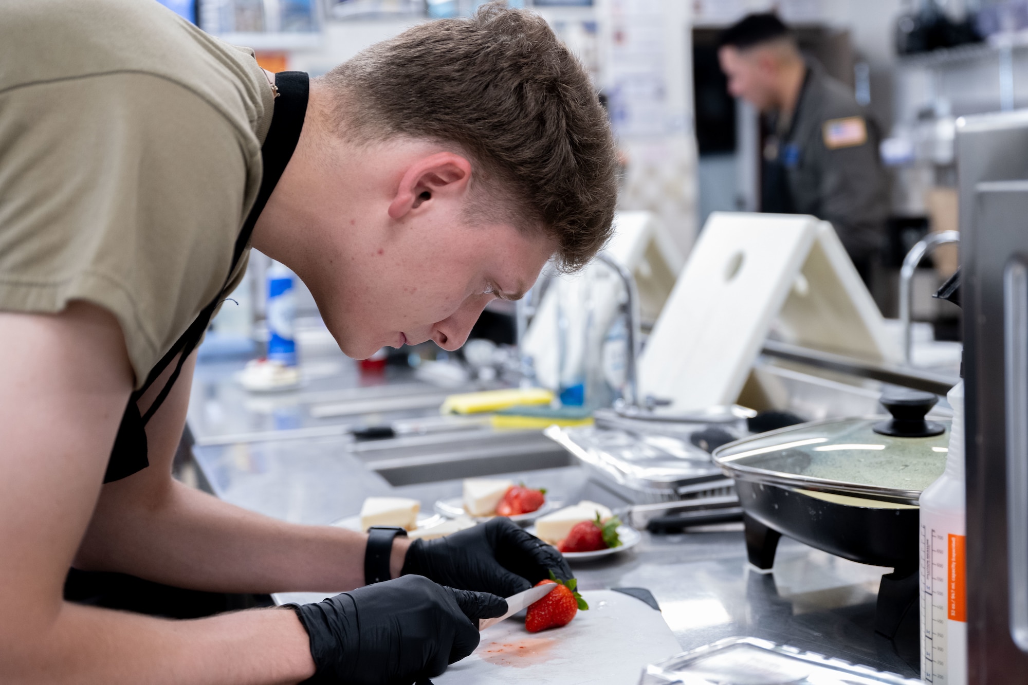An airman slices a strawberry next to a slice of cheesecake.