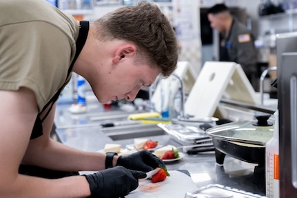 An airman slices a strawberry next to a slice of cheesecake.