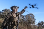 A Marine in tactical gear reaches up towards a drone flying above a forest setting during the day.