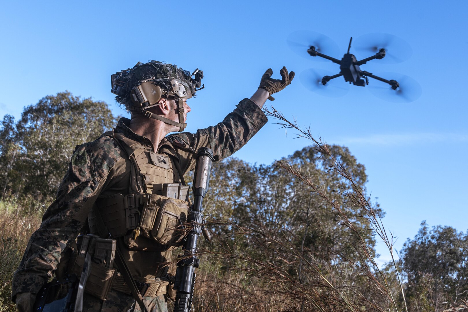 A Marine in tactical gear reaches up towards a drone flying above a forest setting during the day.