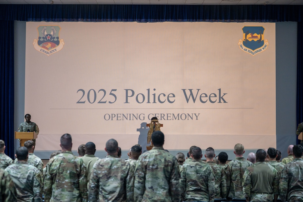 A group of 386th ESFS Airmen stand at attention along the bottom of frame, faced away from the camera toward a projector taking up the remainder of the frame with the title "2025 POLICE WEEK OPENING CEREMONY" and an armor stand holding a vest and helmet in front of it with another Airman at a podium at the left of frame orating the event