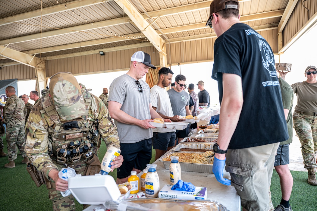 Airmen at the left of the frame form a line to receive barbecue and sides along a table stretched across mid-frame, with 386th ESFS members along the right of frame serving the food