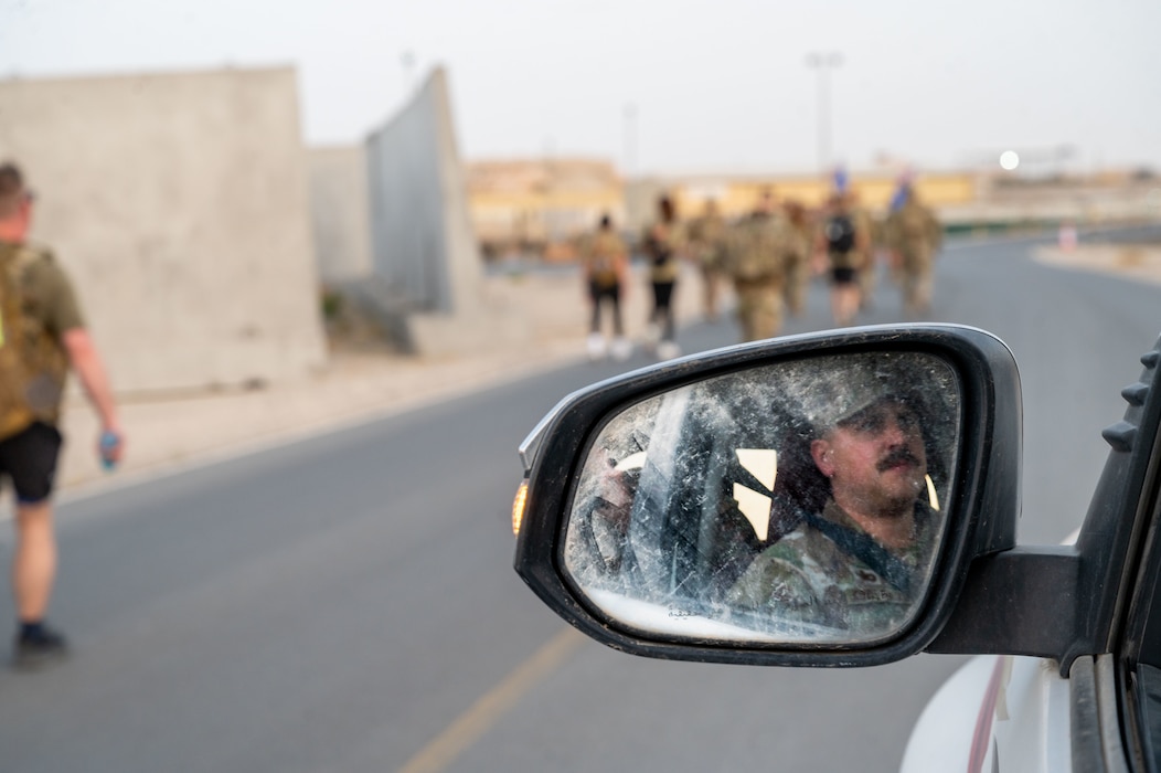 Master Sgt. Austin Hopper is framed within the driver's side car mirror during a patrol along the path of a ruck march with Airmen walking along the side of the path