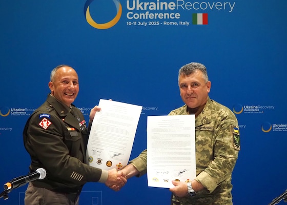 Two men in military uniforms shake hands while displaying signed documents.